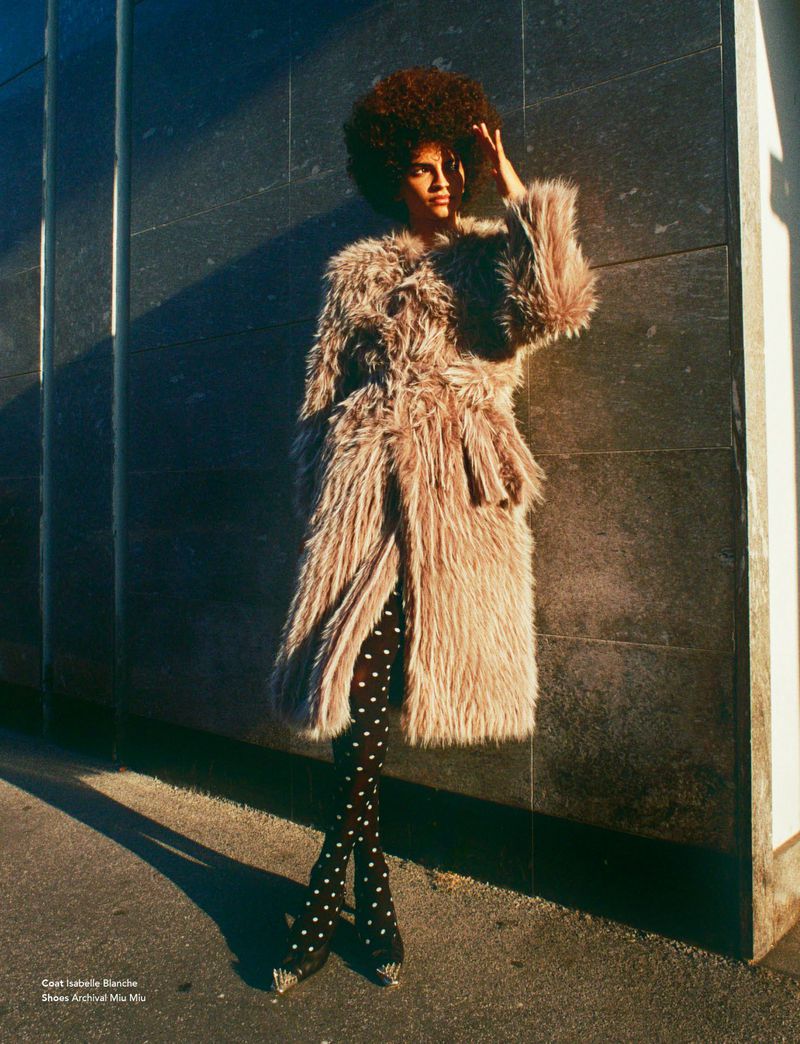 An adult female with an afro poses in a shaggy coat and polka dot tights against a wall, illuminated by sunlight and casting shadows.