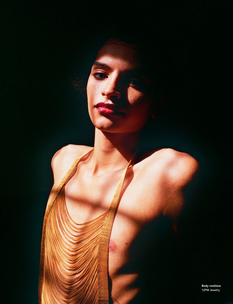 Adult female in a sleeveless golden chainmail top, posing in front of a dark background, light illuminating her face and upper body.