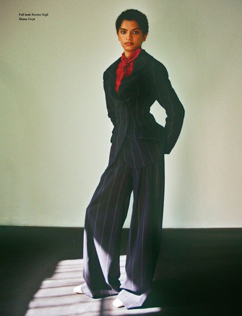 Fashion editorial shot of a woman with short hair, posing in a navy pinstripe suit with a bright red blouse, in front of a neutral wall.