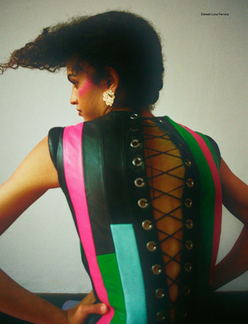 Adult woman wears a colorful corset with elaborate hairstyle against a plain backdrop in a fashion-editorial style photograph.