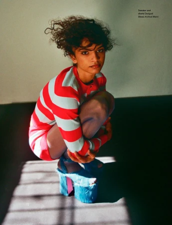 Model with curly hair crouching in a red and white striped sweater with blue platform shoes against a plain wall.