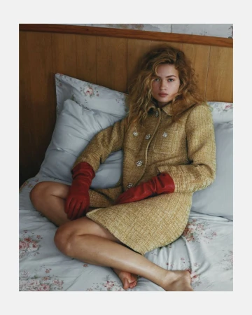 A young woman with blonde curly hair sits cross-legged on a bed wearing a tan tweed suit jacket and skirt, along with red leather gloves.
