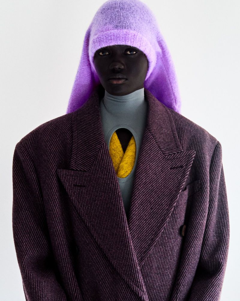 Woman in a purple wool hat and tweed blazer with a gray turtle neck and yellow undershirt, posing against a white background.