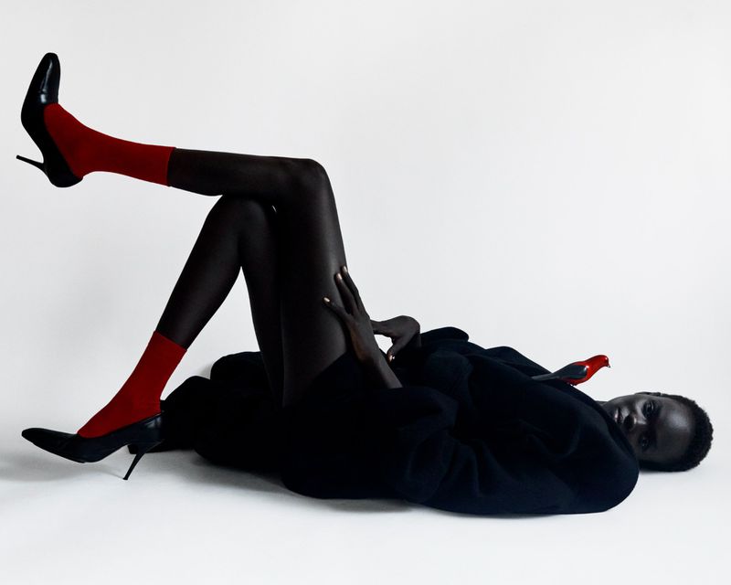 A woman lies on her side wearing black pantyhose, bright red socks and black heels against a white background in a studio fashion shot.