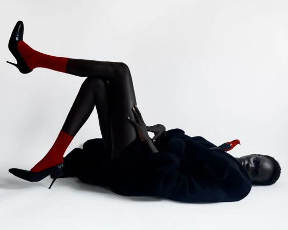 A woman lies on her side wearing black pantyhose, bright red socks and black heels against a white background in a studio fashion shot.