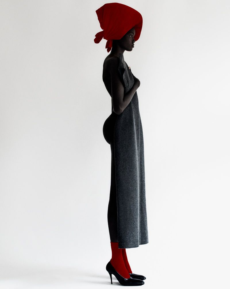 Profile portrait of a Black female fashion model in a grey dress, red stockings, red headscarf and black heels on a white backdrop.