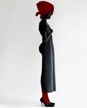 Profile portrait of a Black female fashion model in a grey dress, red stockings, red headscarf and black heels on a white backdrop.