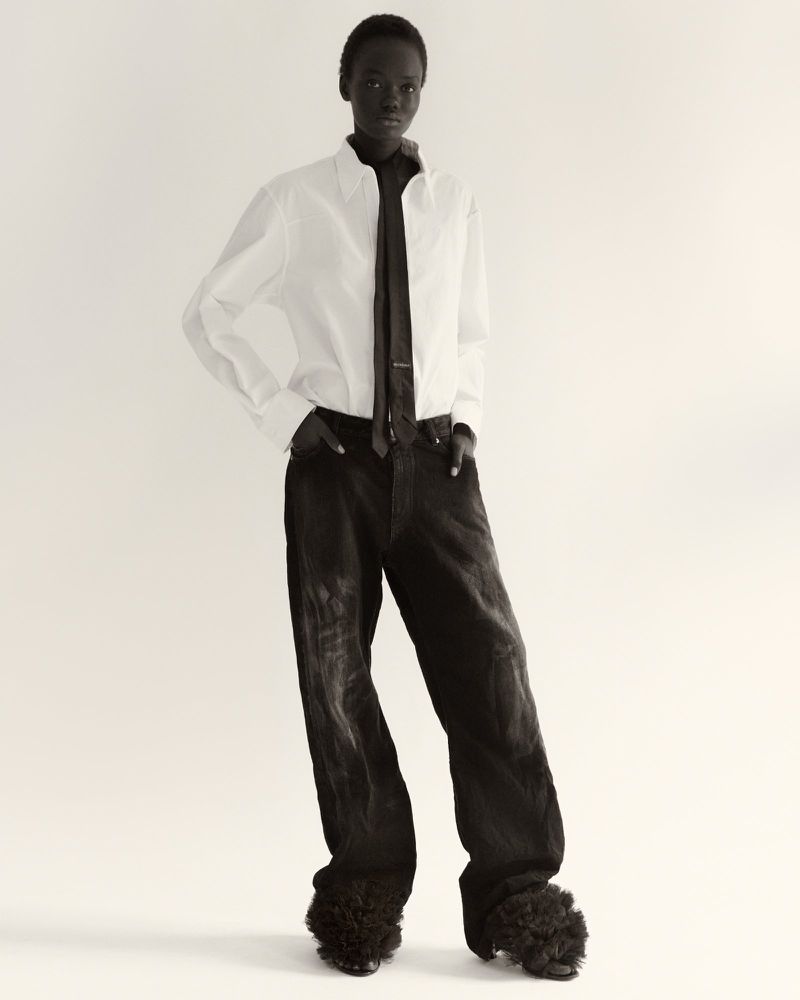 Fashion shot of a Black female model wearing a white shirt, dark tie, baggy black pants, and fur-covered shoes against a plain backdrop.