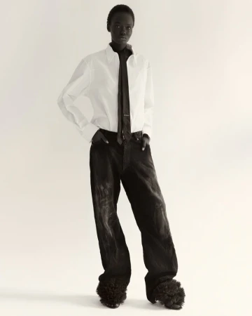 Fashion shot of a Black female model wearing a white shirt, dark tie, baggy black pants, and fur-covered shoes against a plain backdrop.