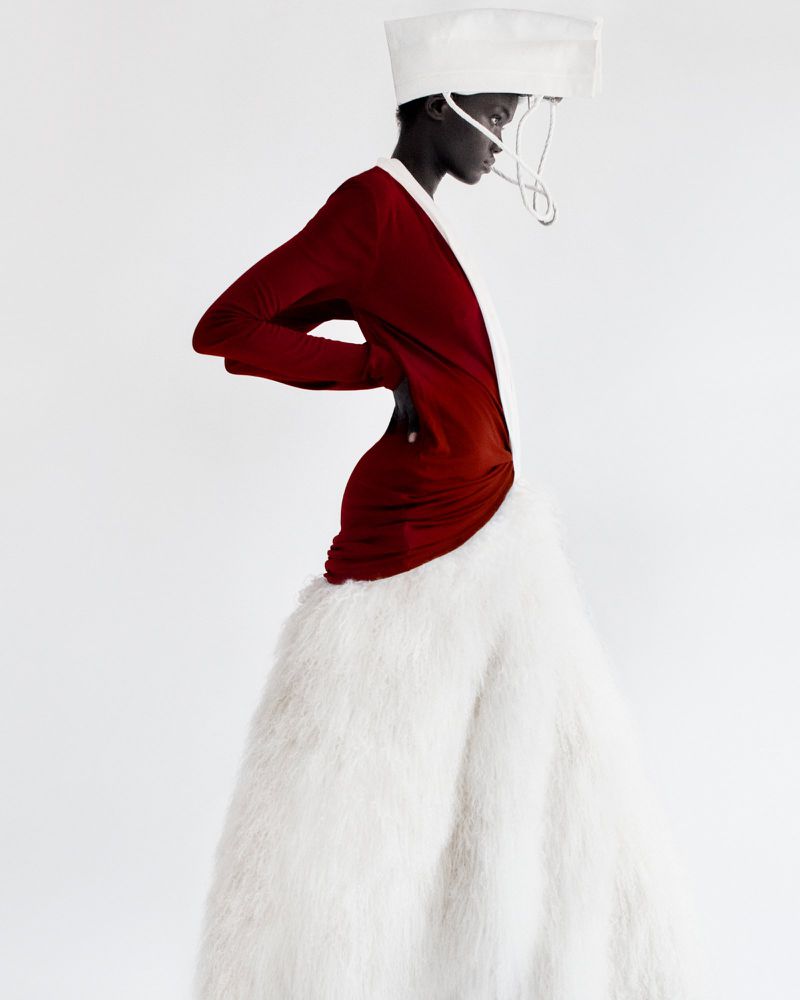 Elegant woman in a dramatic red dress and avant-garde white hat posing against a white background in a fashion forward studio shot.