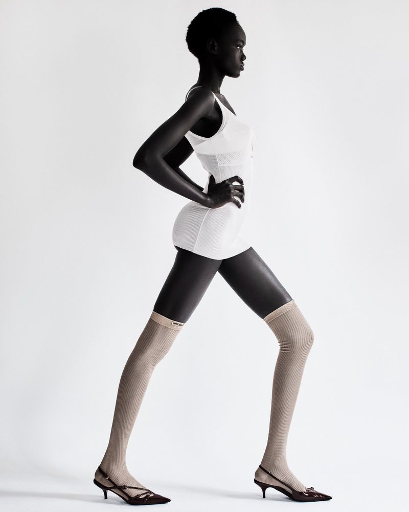 A side-profile studio shot of an adult woman with dark skin wearing a white dress and mesh leg coverings against a plain white background.
