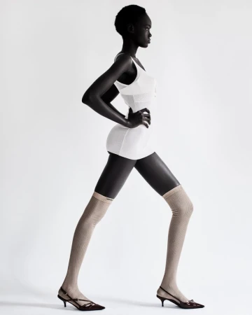 A side-profile studio shot of an adult woman with dark skin wearing a white dress and mesh leg coverings against a plain white background.
