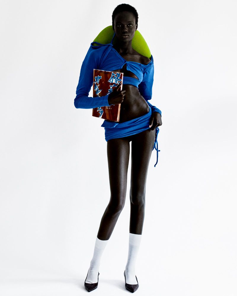 A slender, dark-skinned model in a blue ensemble holds a patterned book in a white studio, showcasing a bold fashion statement.