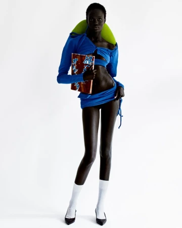 A slender, dark-skinned model in a blue ensemble holds a patterned book in a white studio, showcasing a bold fashion statement.