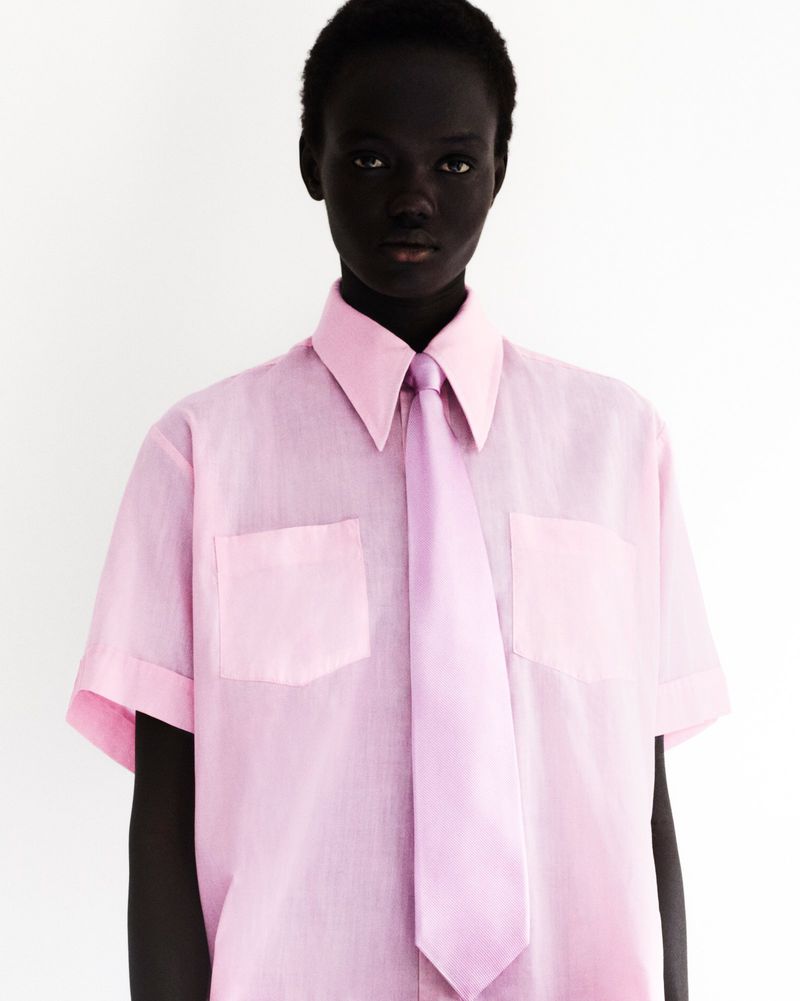 Woman with dark skin poses in a pink short-sleeved shirt with a matching tie against a white background in a studio portrait.