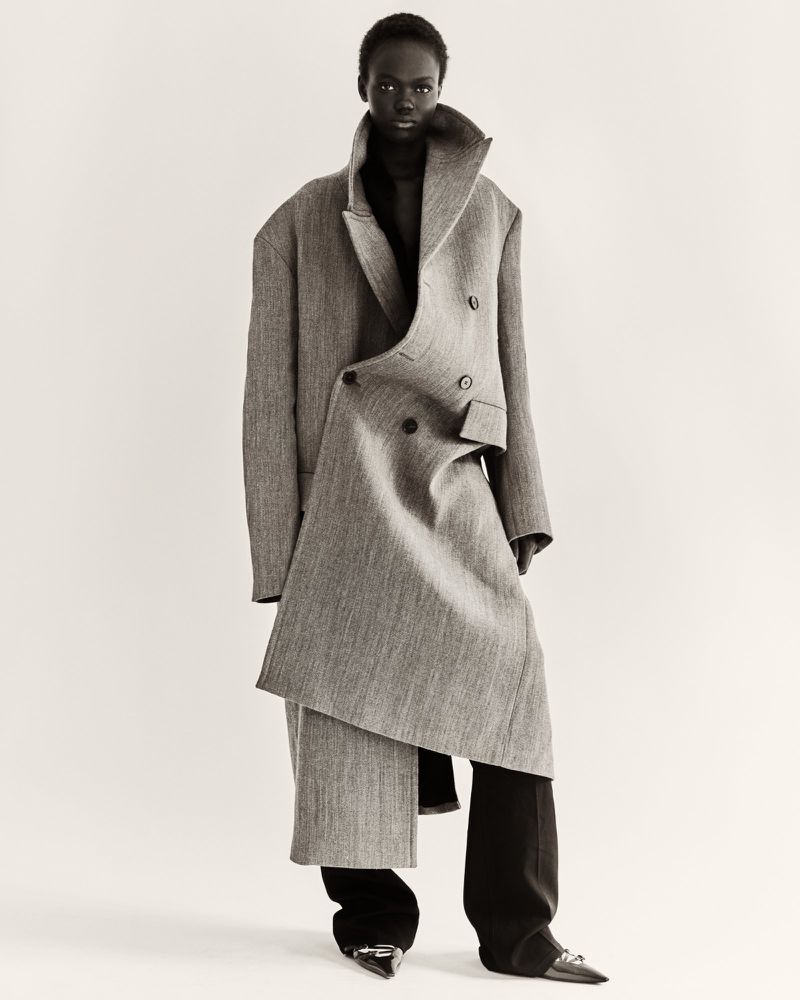 A black female fashion model poses in a studio, wearing a gray wool coat and black pants for a neutral fashion portrait.