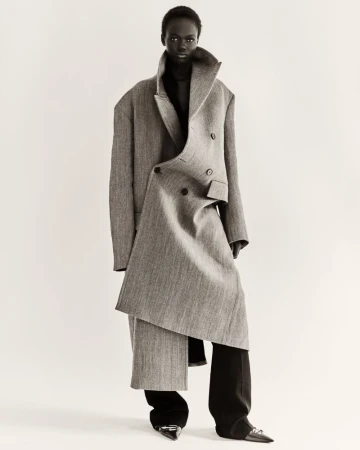 A black female fashion model poses in a studio, wearing a gray wool coat and black pants for a neutral fashion portrait.