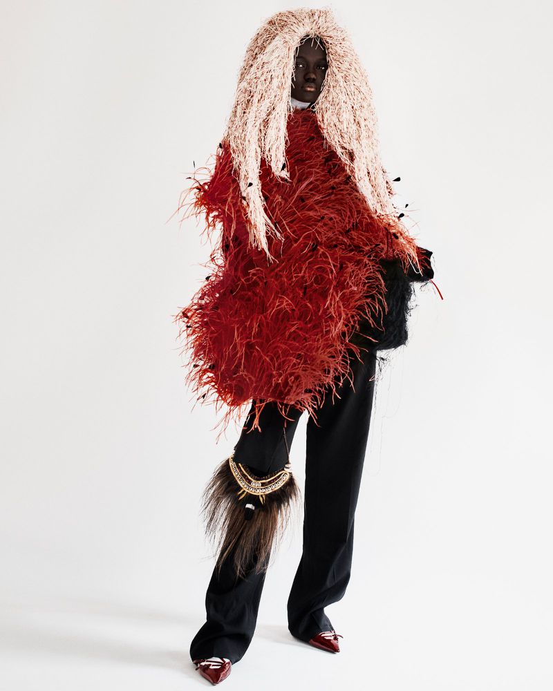 Fashion-forward editorial shot of a woman with an elaborate, feathery headpiece and textured jacket posing in front of a plain white backdrop.