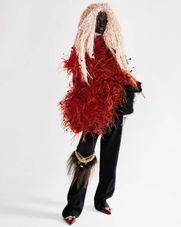 Fashion-forward editorial shot of a woman with an elaborate, feathery headpiece and textured jacket posing in front of a plain white backdrop.