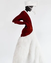Model Achol Kuir in a red top and fluffy white skirt wearing a structured hat against a plain white background