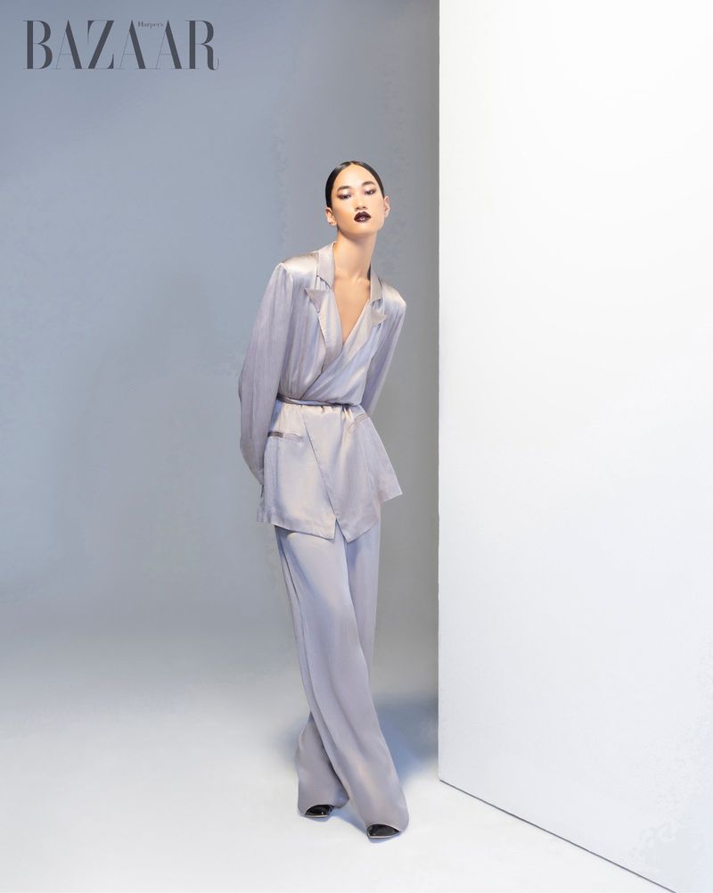 A stylish woman in a light grey, satin suit poses elegantly near a white wall in a studio, presenting a modern and sophisticated look.