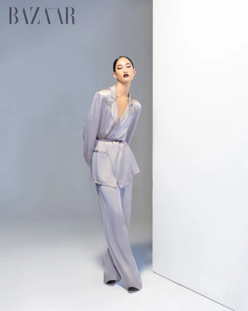A stylish woman in a light grey, satin suit poses elegantly near a white wall in a studio, presenting a modern and sophisticated look.