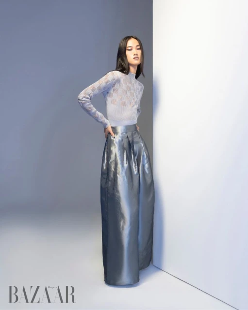 A young adult woman poses in a silver, full skirt and light-blue top against a neutral background in a studio fashion editorial image.