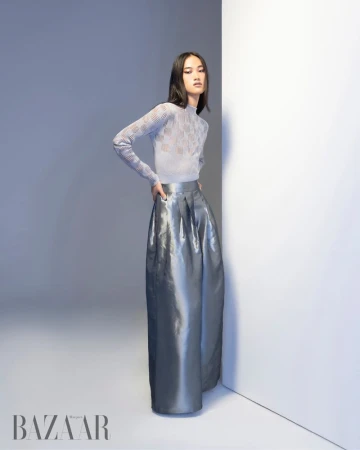 Model in a patterned sheer top and glossy silver wide-leg pants posing against a white studio corner wall.