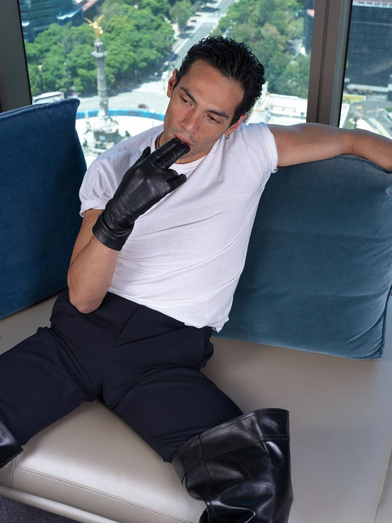 Adult male in black pants, white top, and black gloves poses while sitting on a couch with a blurred urban background.