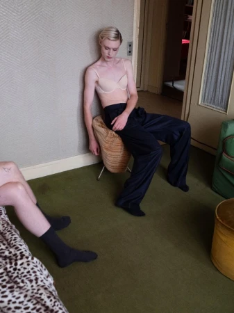 Indoor shot of person with short blonde hair sitting on a stool wearing a bra and dark pants; green carpet in the room.