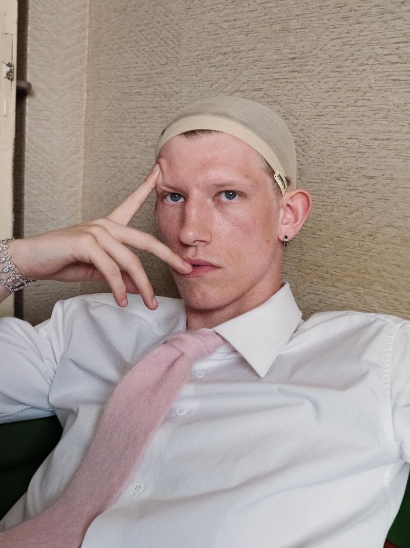 Portrait of a young adult male wearing a wig cap, white dress shirt, and pink tie posing indoors with a neutral facial expression.