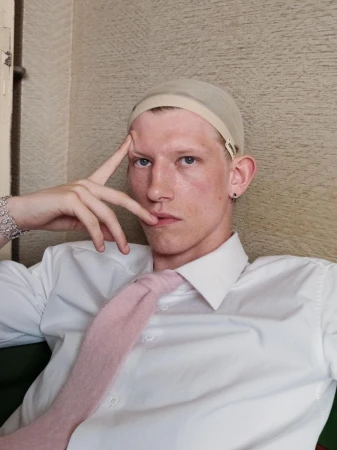 Portrait of a young adult male wearing a wig cap, white dress shirt, and pink tie posing indoors with a neutral facial expression.