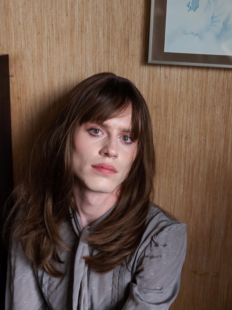 Portrait of a young adult with long brown hair, wearing a grey blouse, posing indoors in front of a patterned wall, with soft, natural lighting.