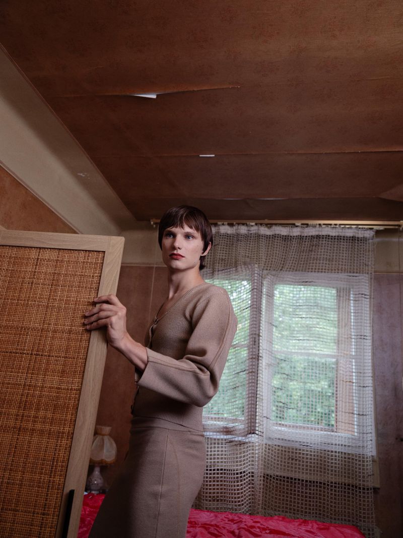 Adult female in a beige outfit stands in a room holding a woven panel; window, bed, and textured walls are in the background.