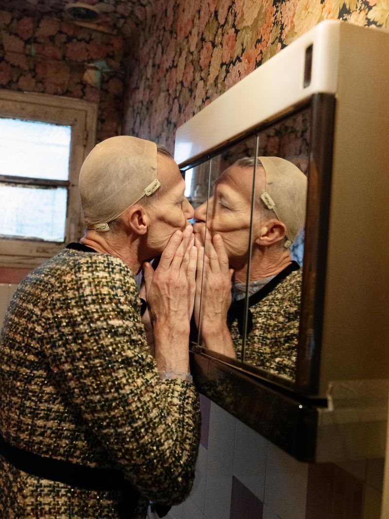 Mature woman with a mesh cap kisses her reflection in a bathroom mirror, wearing a patterned jacket in a cozy interior setting.