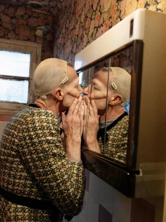Mature woman with a mesh cap kisses her reflection in a bathroom mirror, wearing a patterned jacket in a cozy interior setting.