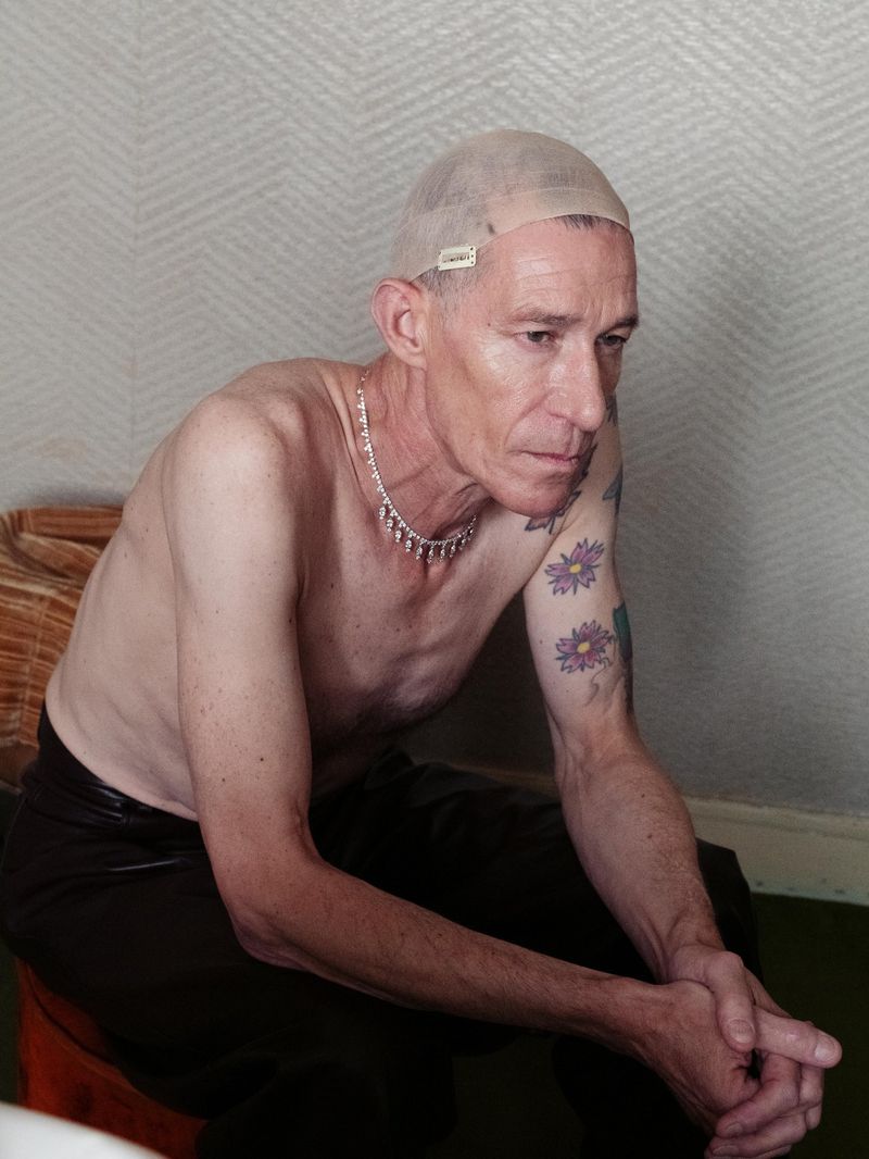 Older man with mesh cap, bare chest, necklace and arm tattoos sits indoors in medium shot.