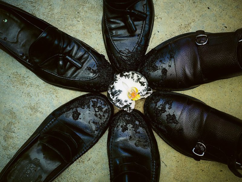 Overhead shot of black leather dress shoes arranged in a circle around a white orchid on a textured gray surface.