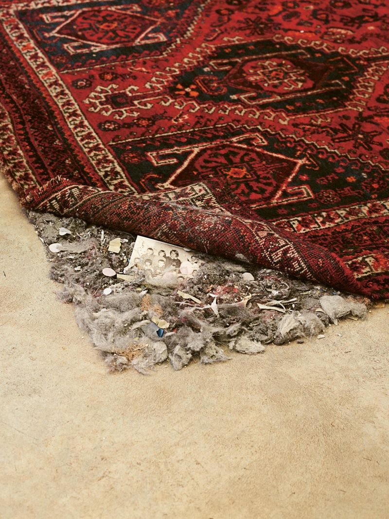 Close-up of a distressed red and black oriental rug showing wear and damage, with exposed backing and debris on a neutral floor.