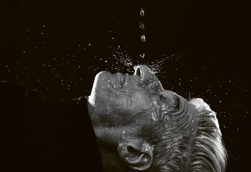 Dramatic black and white studio portrait of an adult with water droplets falling on their face, looking upward against a dark background.