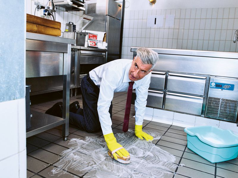 Businessman in office attire kneeling to clean a tiled floor with a sponge and soapy water, featuring cleaning supplies in a commercial kitchen.