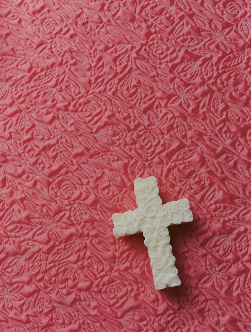 Close-up studio shot of a white, decorative cross with floral patterns on a pink, textured, floral-patterned fabric background.