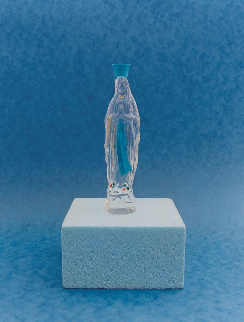 Clear plastic Virgin Mary figurine with blue accents sitting on a light blue cube against a blue backdrop in a studio setting.