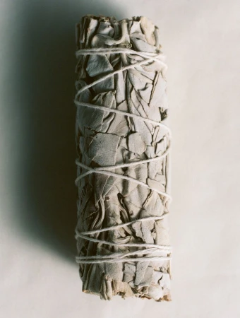 A vertical bundle of dried white sage leaves wrapped tightly with white twine against an off-white background.