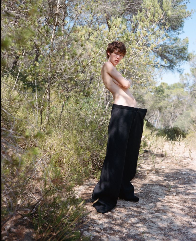 Young adult poses topless in oversized black pants amid lush green foliage. Sunlight filters through trees in this outdoor portrait.