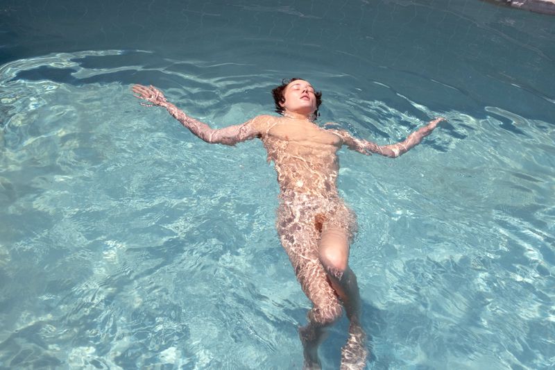 Woman floats in light blue swimming pool water with her arms outstretched. The scene is brightly lit and shows a serene moment.