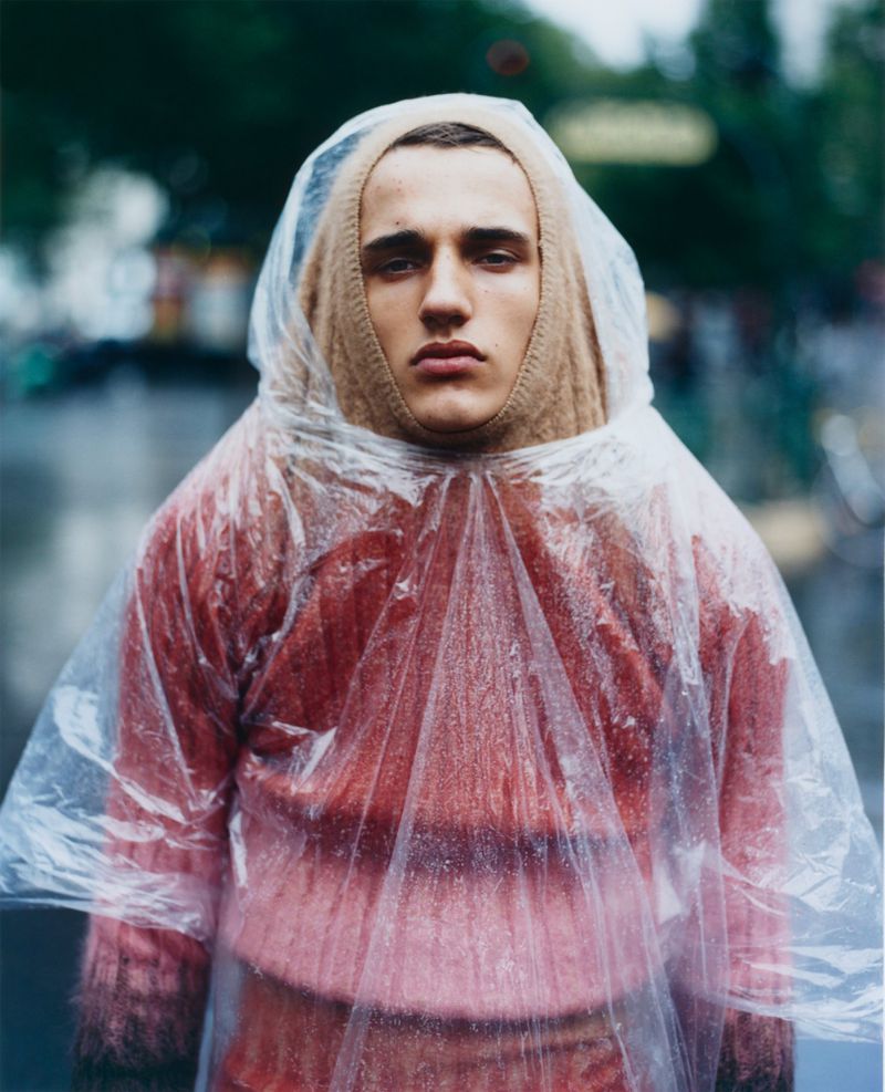 A young adult man wearing a clear poncho and knit balaclava poses in an eye-level portrait shot with a blurred outdoor urban background.