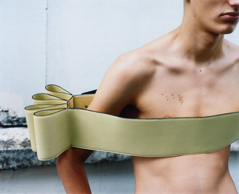 Male fashion model with bare chest poses in front of a concrete wall, wearing a structured, light-green, shoulder-hugging garment.