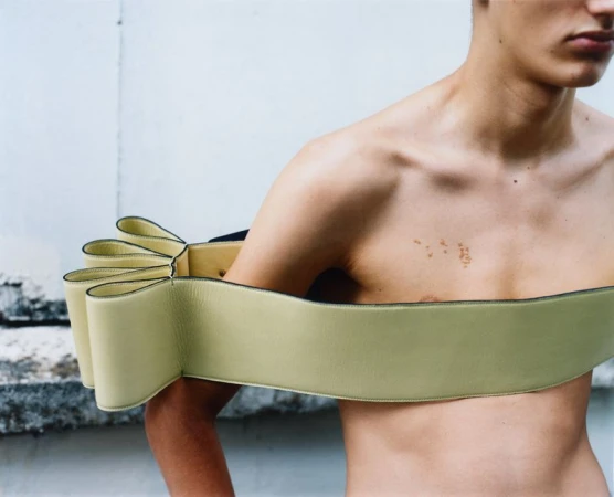 Male fashion model with bare chest poses in front of a concrete wall, wearing a structured, light-green, shoulder-hugging garment.