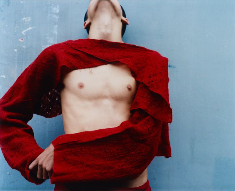 Torso of a slender adult male in red knitwear posing against a pale blue wall, angled upward showing his neck and abdomen.